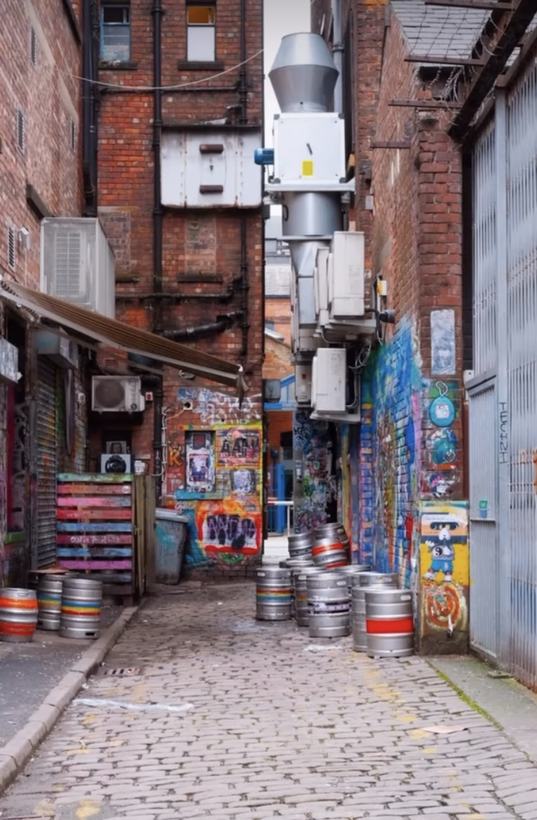 Fotografi: smal bakgate med brostein, ølfat, teglvegger, ventilasjonsrør og fargerik graffiti.