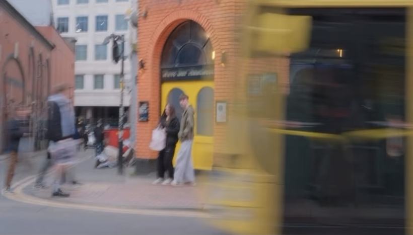 Fotografi: et gatehjørne med oransje murbygning, delvis dekket av en gul, uskarp buss i fart.