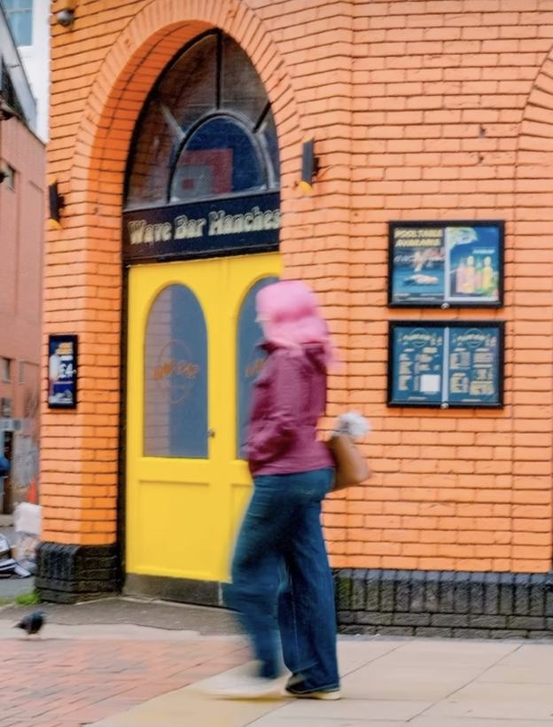 Fotografi: en person i rosa hodeplagg går forbi en knallgul dør i en oransje murvegg, i bevegelsesuskarphet.